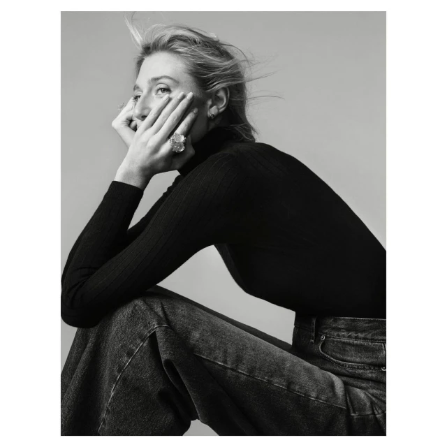 Monochrome portrait of an adult woman with blonde hair wearing a black turtleneck sweater and blue jeans, seated in a photo studio.