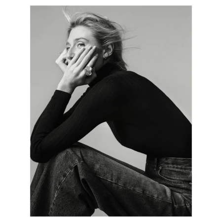 Monochrome portrait of an adult woman with blonde hair wearing a black turtleneck sweater and blue jeans, seated in a photo studio.