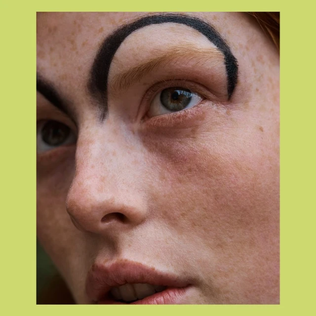 Close-up portrait of a fair-skinned adult woman with distinctive makeup, featuring a dramatic, dark eyebrow design and green eyes.