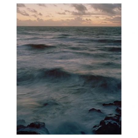 Ocean waves crashing against rocks under a cloudy sky, captured with a long exposure to create a blurred water effect.