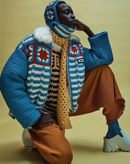 A person wearing a crocheted jacket, hat, and scarf poses in a studio with a yellow background, sitting with one knee bent.