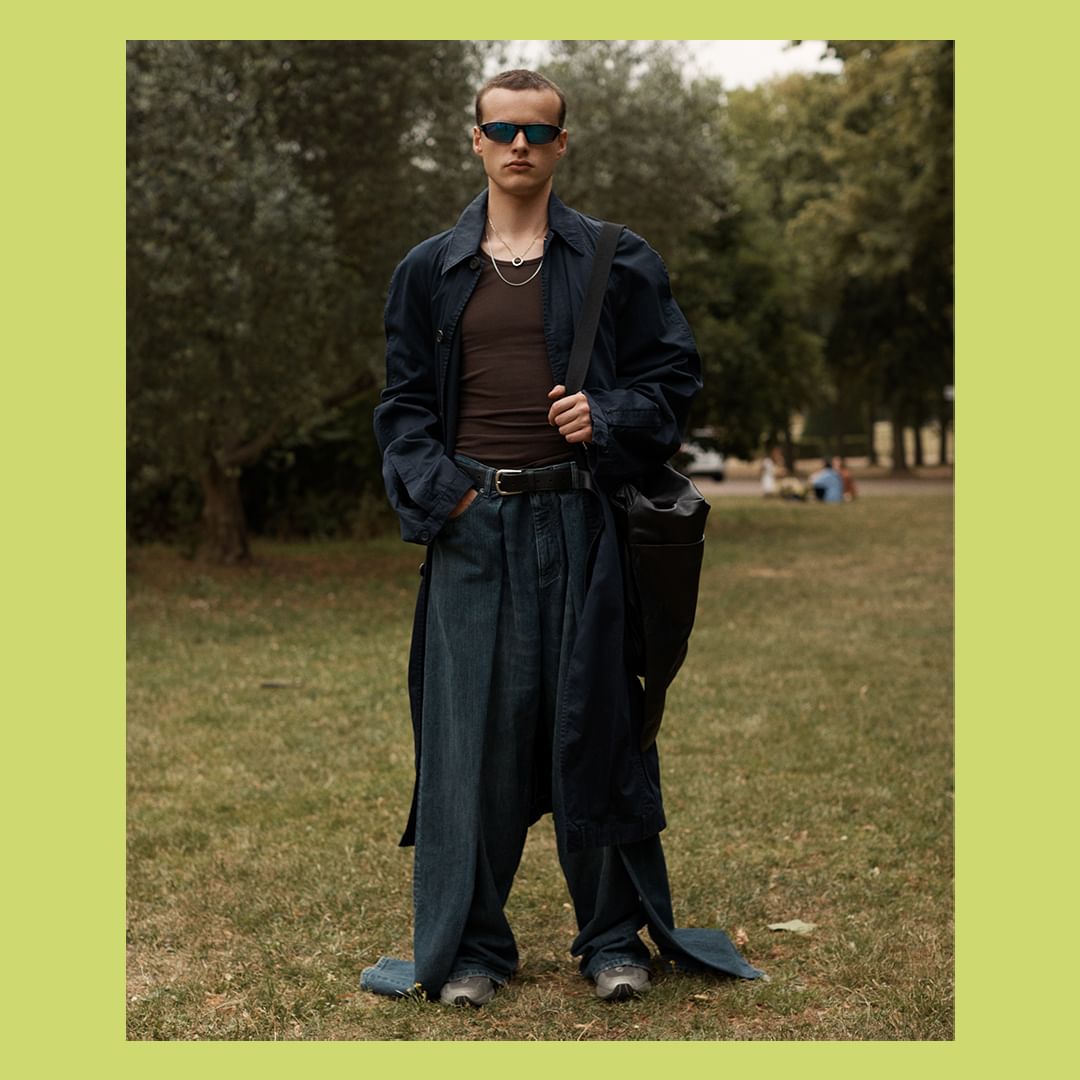 Full shot of a slender adult man in oversized jeans, sunglasses, and a long coat standing on a grassy lawn with trees in the background.