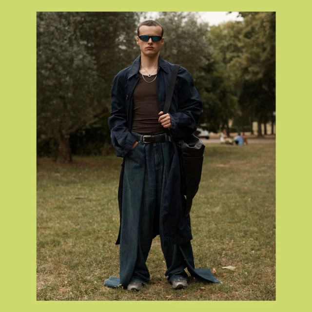 Full shot of a slender adult man in oversized jeans, sunglasses, and a long coat standing on a grassy lawn with trees in the background.