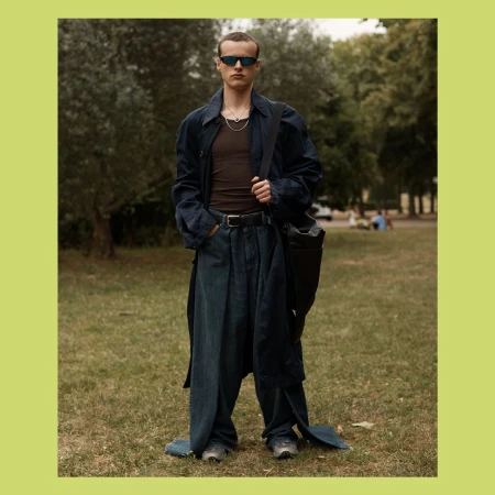 Full shot of a slender adult man in oversized jeans, sunglasses, and a long coat standing on a grassy lawn with trees in the background.
