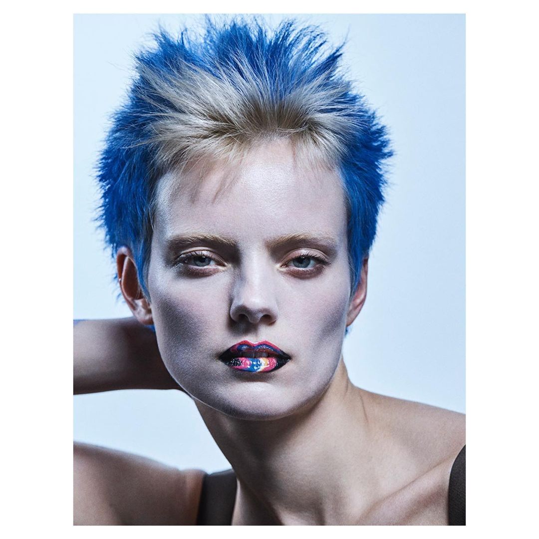 Close-up of a model with short blue and blonde hair and colorful lips, in a beauty shot against a light blue background in studio.