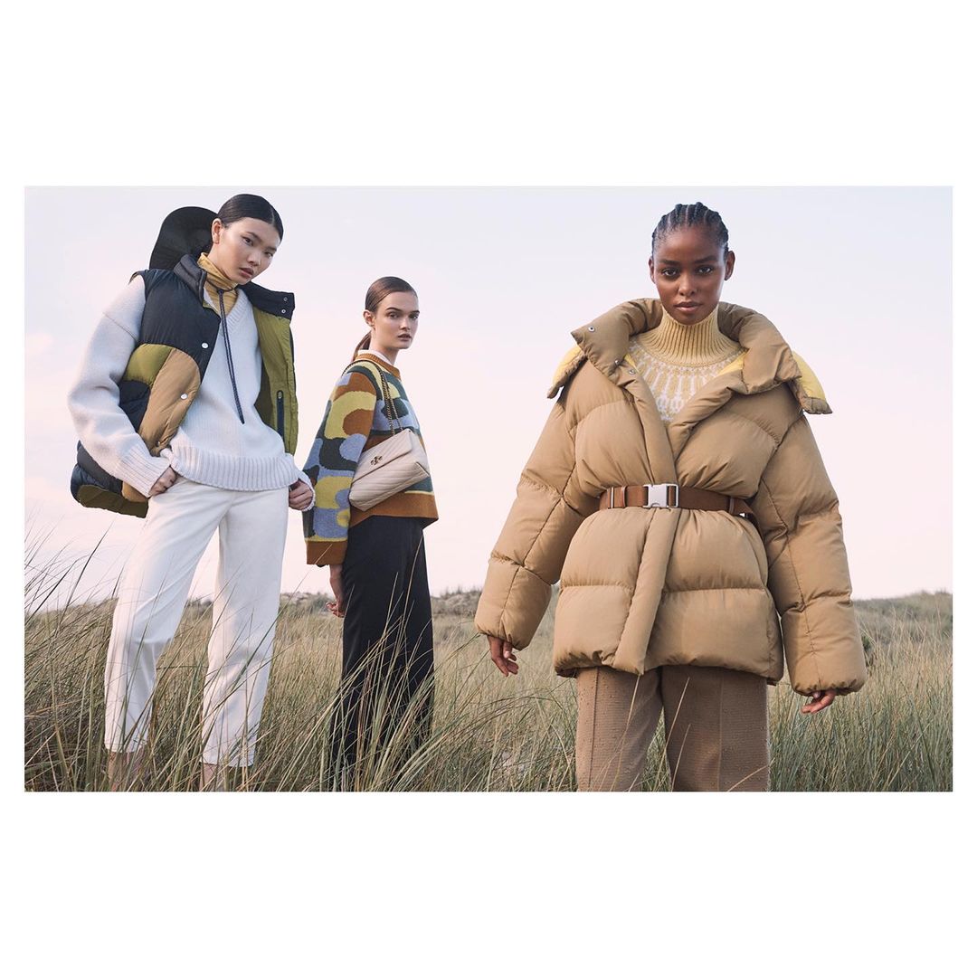 Three women in fashionable outerwear stand in a field of tall grass. The focus is on their clothing and the neutral color palette.