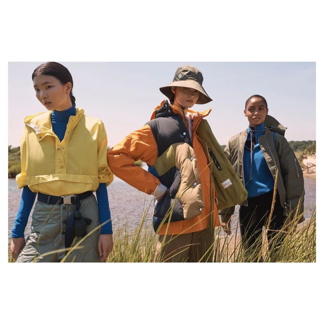 Three women stand outside in layered jackets and pants, showcasing fashion against a backdrop of grass and water on a bright day.