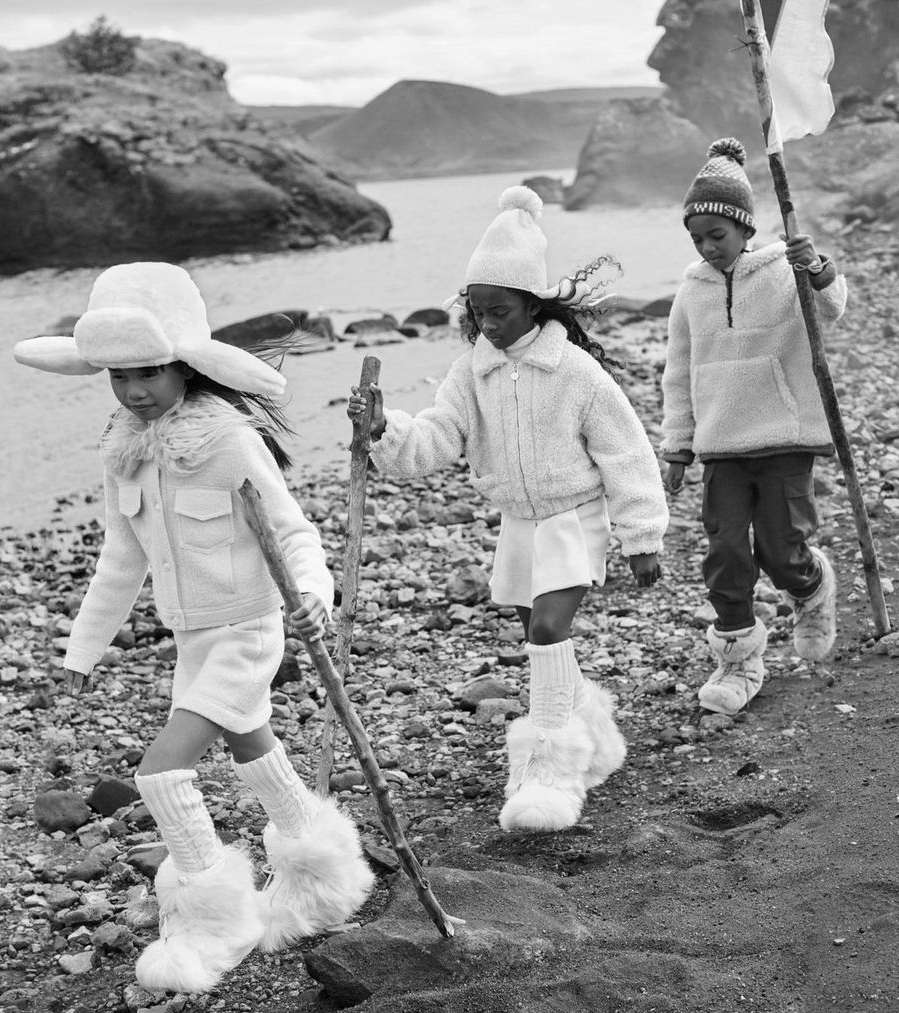 Three children wearing winter clothing walk on a rocky beach near the ocean. The monochrome shot features winter fashion and landscape.