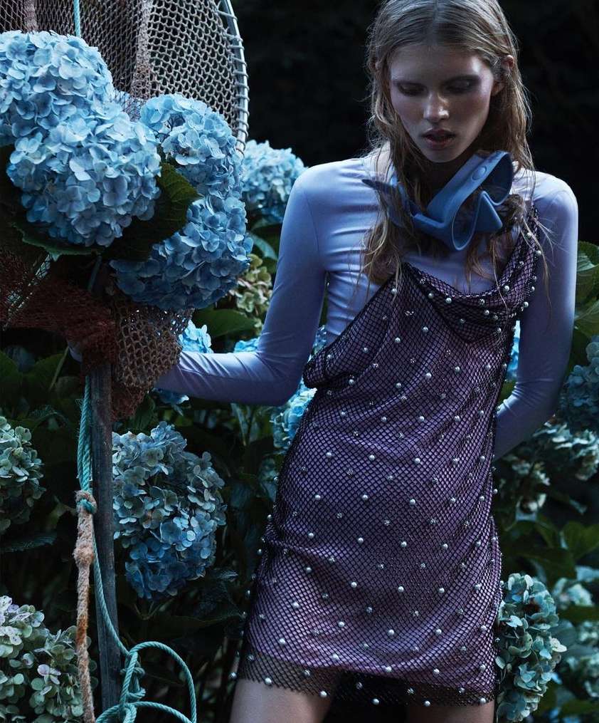 Adult woman in a slip dress poses with blue hydrangeas and netting outdoors in a fashion shoot; dark and moody lighting