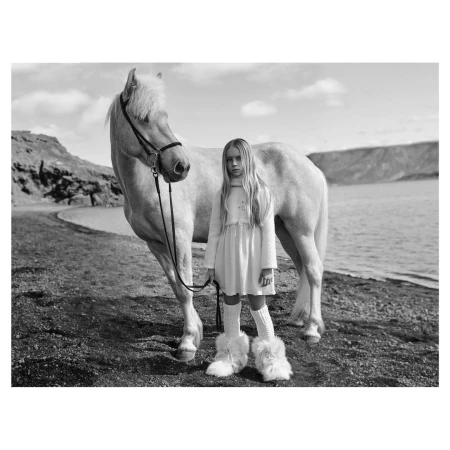 Young girl standing on a pebbled beach next to a white horse holding its reins, wearing a pale dress and furry boots.