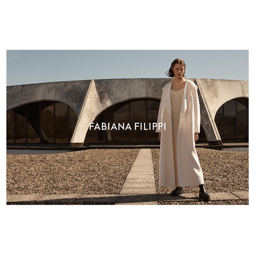 An adult woman stands outdoors in a light-colored coat, dress, and black boots in front of a unique circular concrete building.  
