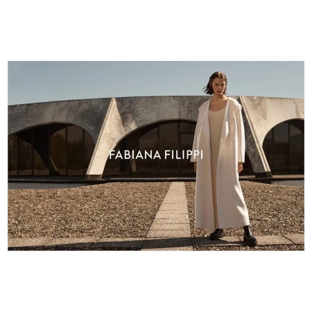 An adult woman stands outdoors in a light-colored coat, dress, and black boots in front of a unique circular concrete building.  