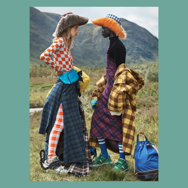 Two women in eye-catching hats and colorful outfits face each other outdoors amidst a hilly, grassy landscape, creating a visually striking editorial shot.