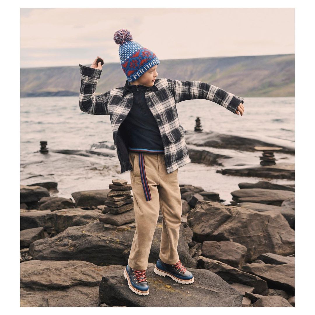 Boy throwing stone on rocky beach with mountains and ocean background. Wearing plaid jacket, hat, pants and boots in outdoor setting.