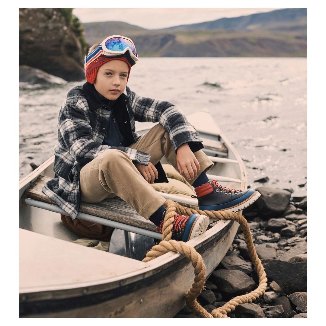 Boy wearing ski goggles and plaid shirt sits in a canoe on a lake with forested mountains in the background in an outdoor setting.