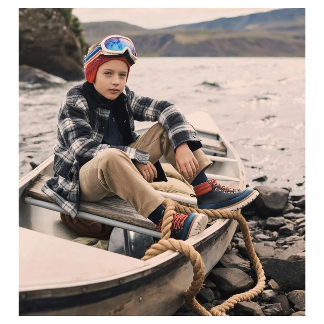 Boy wearing ski goggles and plaid shirt sits in a canoe on a lake with forested mountains in the background in an outdoor setting.