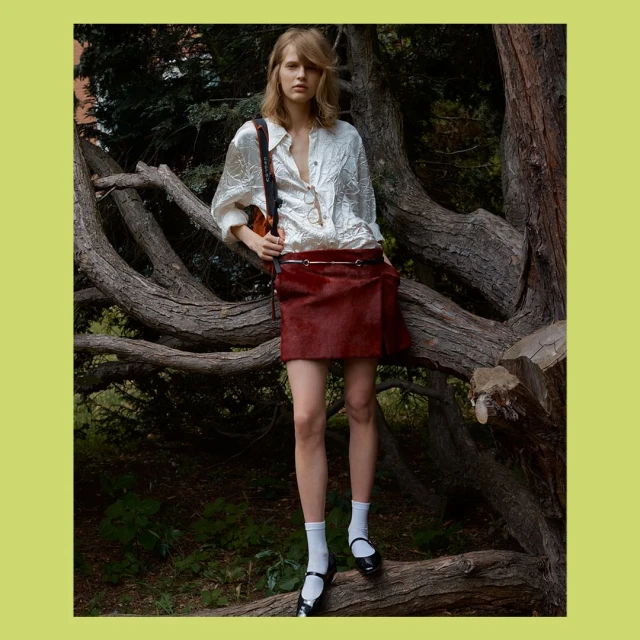 Young woman stands on a fallen tree in a park wearing a white embroidered blouse and reddish brown miniskirt with black Mary Janes.