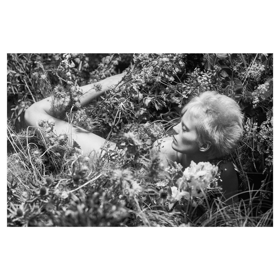 Black and white portrait of a woman reclining amidst dense garden foliage and wildflowers, captured by Josh Olins.