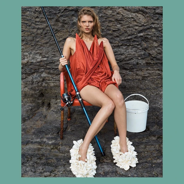 Fashion portrait of a blond woman in an orange dress sitting in a chair with a fishing pole, wearing floral shoes, near a bucket and rocks.
