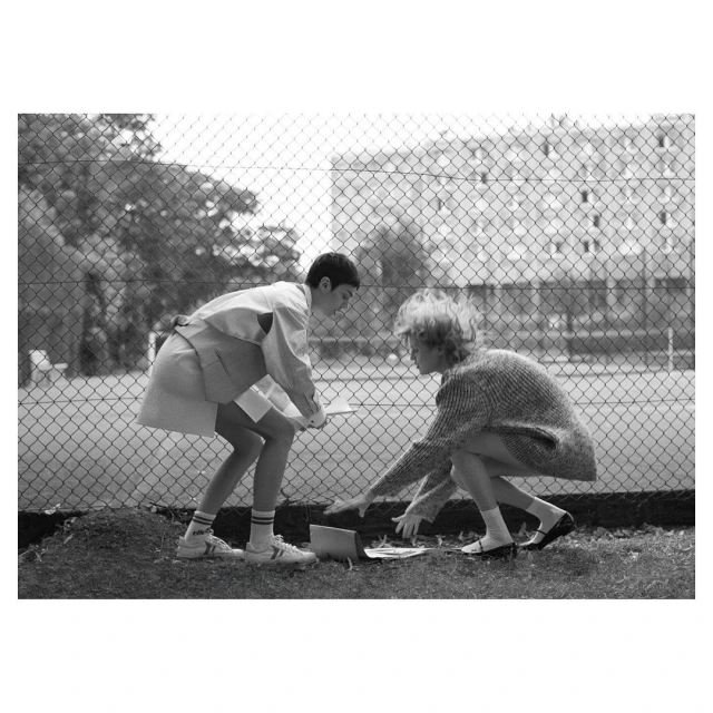 Two young people in an outdoor setting, inspecting items on the ground near a chain-link fence and a building in the background.