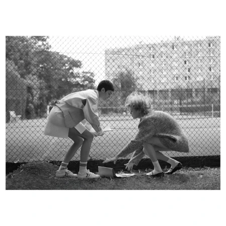 Two young people in an outdoor setting, inspecting items on the ground near a chain-link fence and a building in the background.