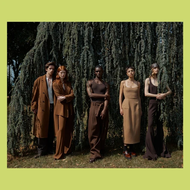 Group of five fashion models pose in an outdoor setting, wearing earth-toned clothing, standing in front of a large evergreen tree.