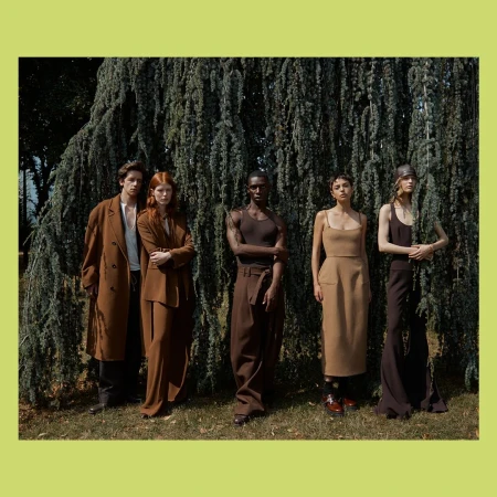 Group of five fashion models pose in an outdoor setting, wearing earth-toned clothing, standing in front of a large evergreen tree.