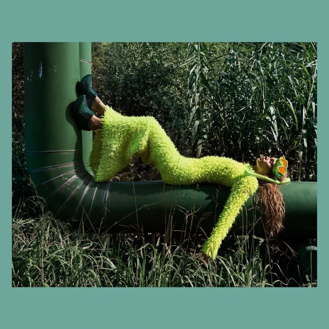 Fashion shot of a woman in a lime green fuzzy suit lying on a green pipe outdoors surrounded by tall grass and greenery.