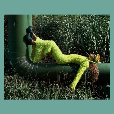Fashion shot of a woman in a lime green fuzzy suit lying on a green pipe outdoors surrounded by tall grass and greenery.