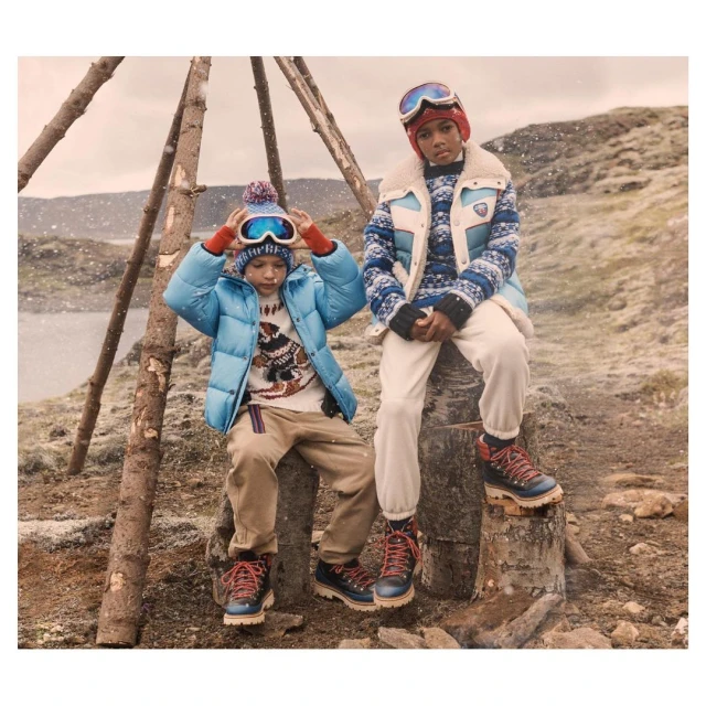 Two children in winter clothing sit on tree stumps in a snowy outdoor scene, wearing puffer jackets, sweaters, ski goggles, and winter boots.