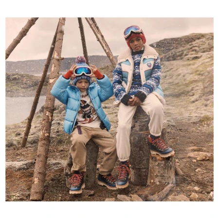 Two children in winter clothing sit on tree stumps in a snowy outdoor scene, wearing puffer jackets, sweaters, ski goggles, and winter boots.