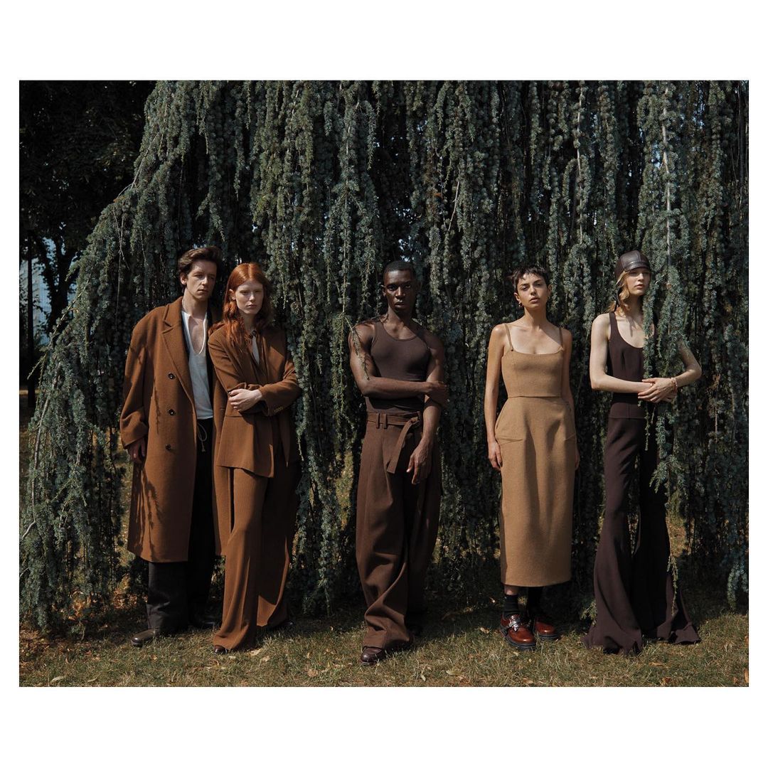 Group of five fashion models in earth-tone vintage clothing posing outdoors in front of a large, drooping evergreen tree in a fashion editorial shoot.