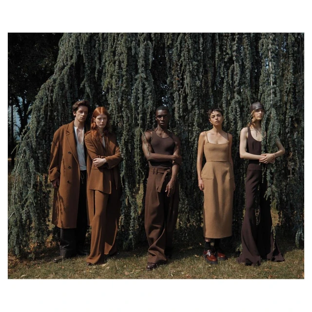 Group of five fashion models in earth-tone vintage clothing posing outdoors in front of a large, drooping evergreen tree in a fashion editorial shoot.