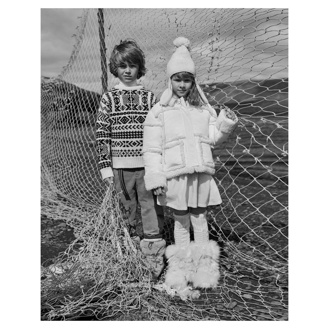 Black and white portrait of a boy in a patterned sweater and a girl in a white coat and hat standing by a fishing net.