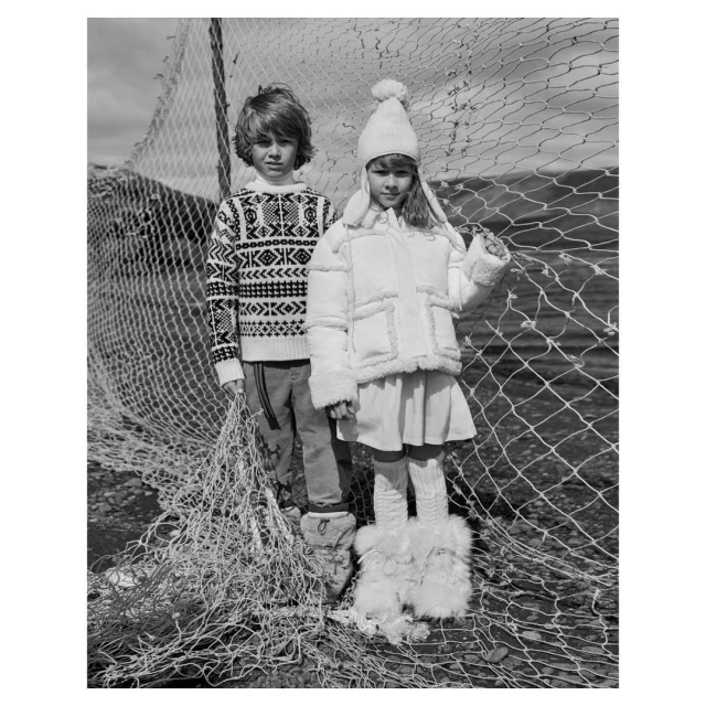 Black and white portrait of a boy in a patterned sweater and a girl in a white coat and hat standing by a fishing net.