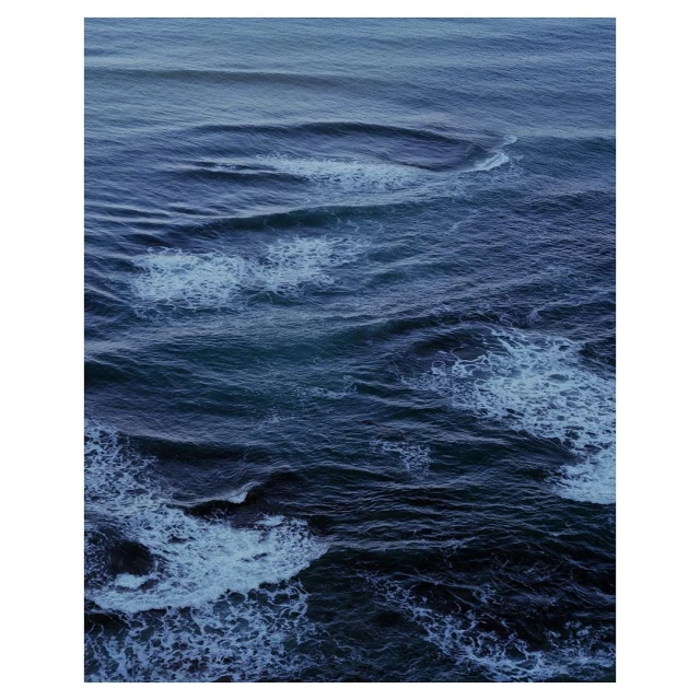 An aerial view captures the ocean's expanse, showcasing waves and foam patterns in a range of blues from dark navy to light turquoise.