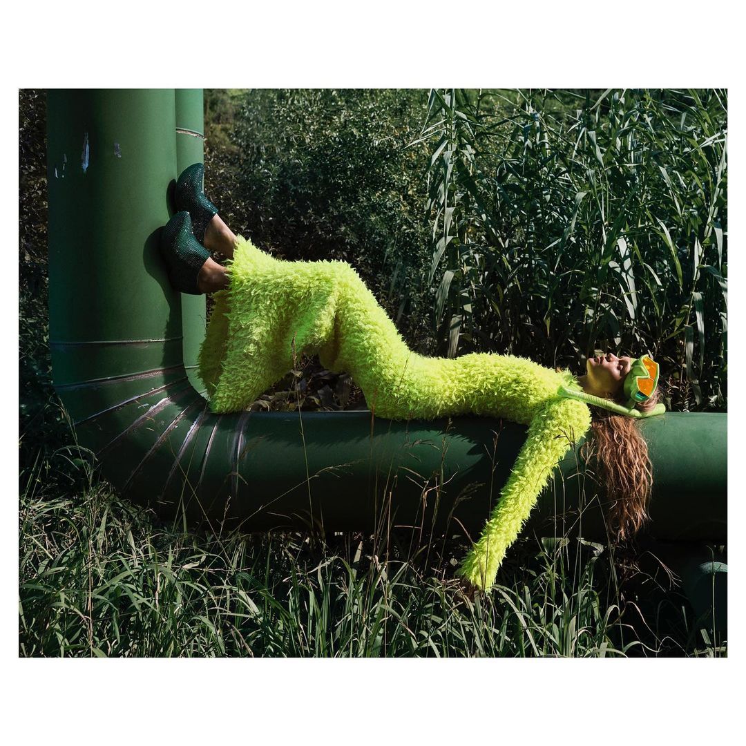 Woman in a fuzzy green suit lying on a green pipe outdoors surrounded by tall grass and plants in a fashion editorial setting.