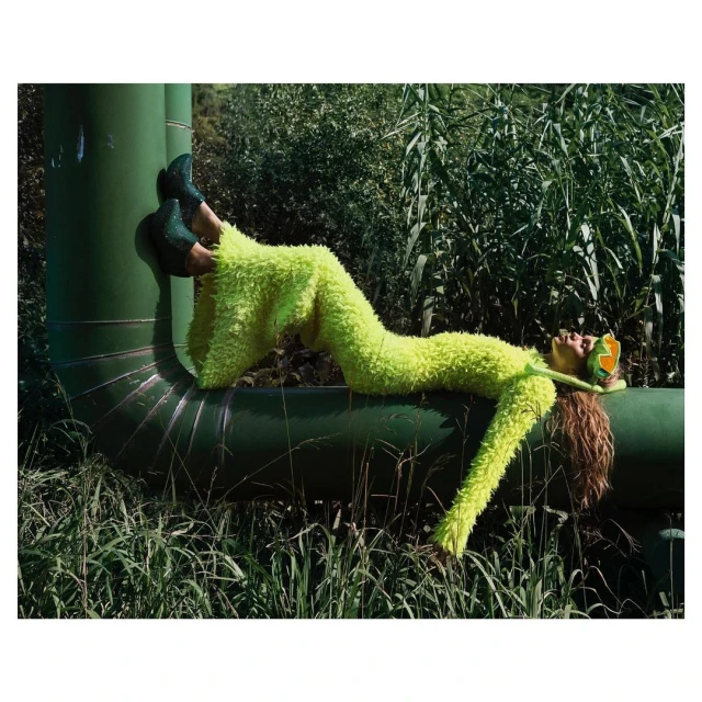 Woman in a fuzzy green suit lying on a green pipe outdoors surrounded by tall grass and plants in a fashion editorial setting.
