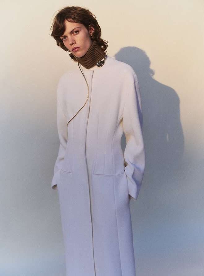 Fashionable adult female model posing in a cream-colored coat with a brown turtleneck against a gradient studio backdrop.