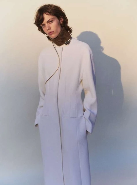 Fashionable adult female model posing in a cream-colored coat with a brown turtleneck against a gradient studio backdrop. 