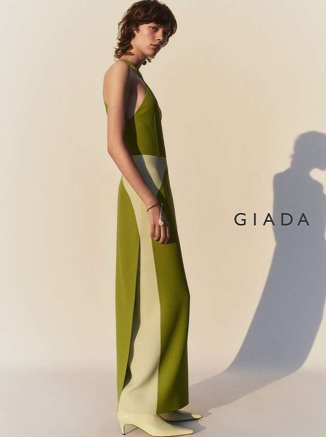 An adult female fashion model poses indoors wearing a two-tone halter dress with green and beige vertical color blocking and kitten heels.