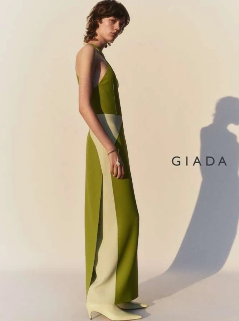 An adult female fashion model poses indoors wearing a two-tone halter dress with green and beige vertical color blocking and kitten heels.