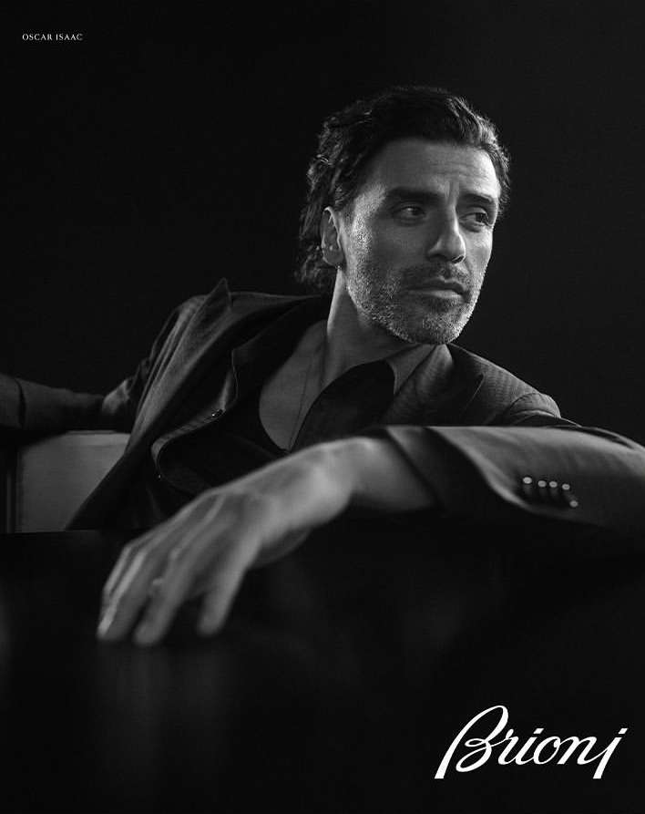 Black and white portrait of Oscar Isaac wearing a suit jacket, gazing slightly off-camera, with the Brioni logo in the corner.
