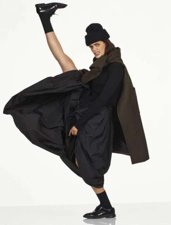 Model in black beanie and dark clothing kicking leg high wearing black leather shoes against white background