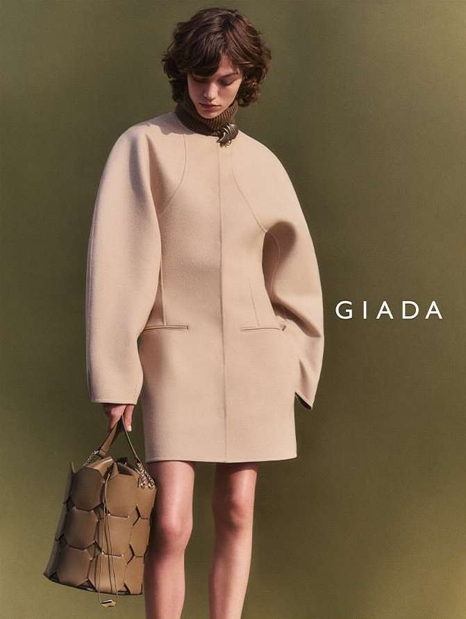 Fashion shot of a woman in a beige coat dress and carrying a tan handbag, posing against a muted olive green backdrop indoors.