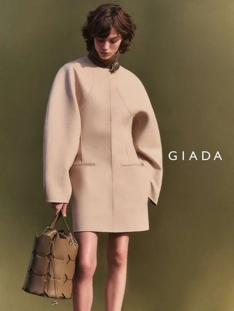 Fashion shot of a woman in a beige coat dress and carrying a tan handbag, posing against a muted olive green backdrop indoors.