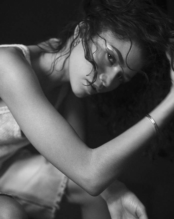 Monochrome portrait of a young woman with curly hair, gazing downwards in a dimly lit environment, showcasing her bare arm with a bracelet.