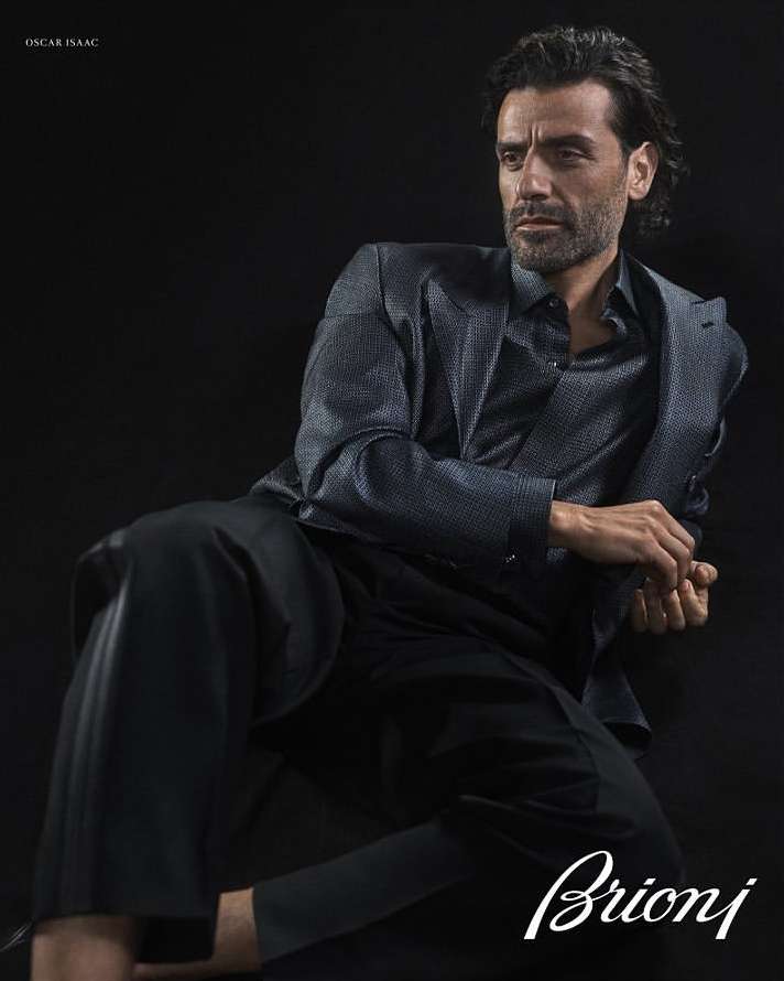 Portrait of Oscar Isaac in a suit jacket and dress shirt against a black background in a studio fashion shot. Man is posed.