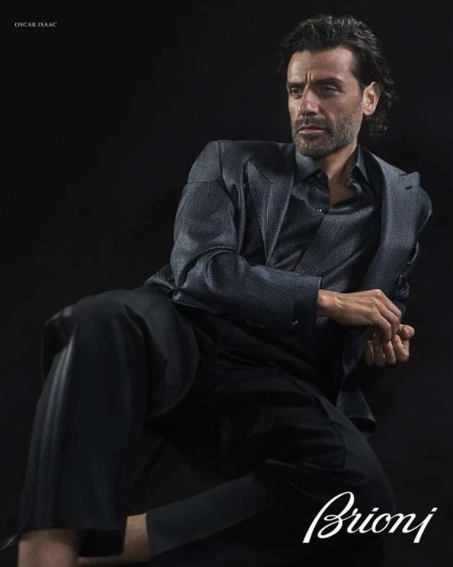 Portrait of Oscar Isaac in a suit jacket and dress shirt against a black background in a studio fashion shot. Man is posed.