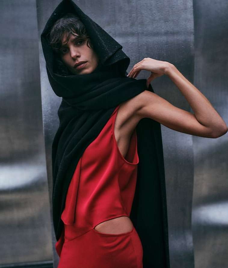Adult female model posing in a sleeveless red dress with cutout detail and a black hooded cloak against a grey metallic background.